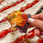 A hand with striped red and white nail art holds a blue corn chip topped with shredded cheese, lettuce, salsa, and a cherry tomato slice. In the background is a layered dip with sour cream, cherry tomatoes, and possibly guacamole.