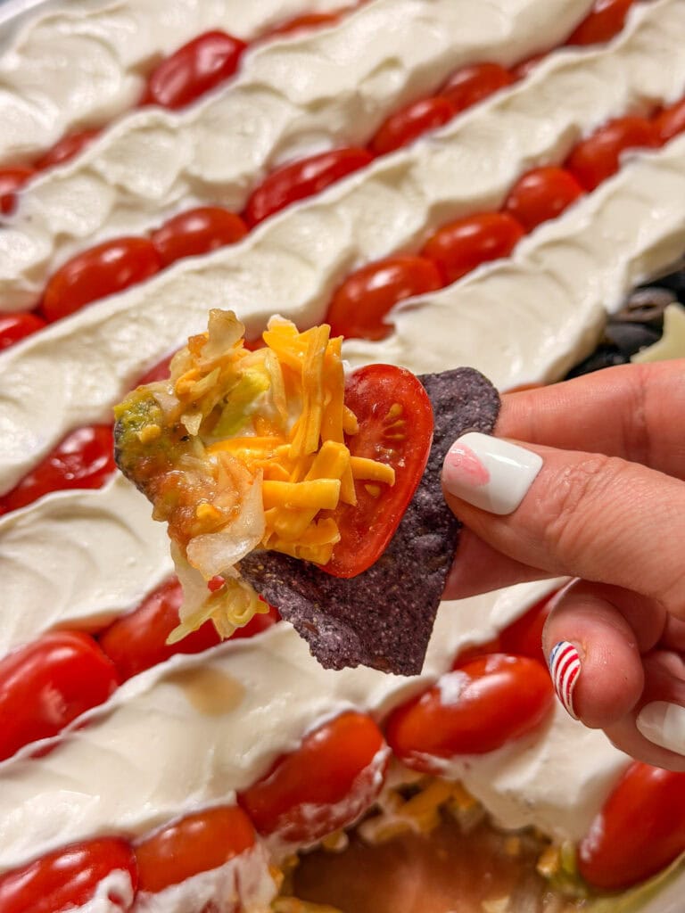 A hand with striped red and white nail art holds a blue corn chip topped with shredded cheese, lettuce, salsa, and a cherry tomato slice. In the background is a layered dip with sour cream, cherry tomatoes, and possibly guacamole.