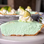 A slice of light green key lime pie with a graham cracker crust, topped with whipped cream and a lime wedge, sits on a white plate. The rest of the pie is visible in the background.