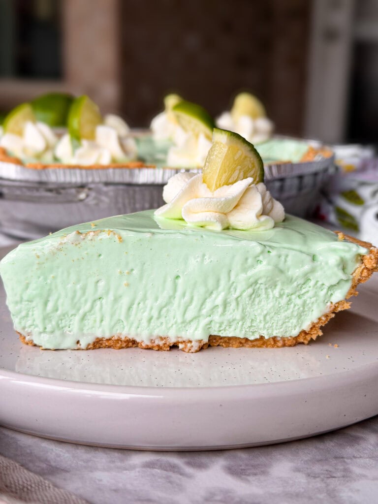 A slice of light green key lime pie with a graham cracker crust, topped with whipped cream and a lime wedge, sits on a white plate. The rest of the pie is visible in the background.