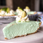 A slice of creamy light green pie with a graham cracker crust, topped with whipped cream and a small lime wedge, sits on a white plate. The whole pie is visible in the blurred background.