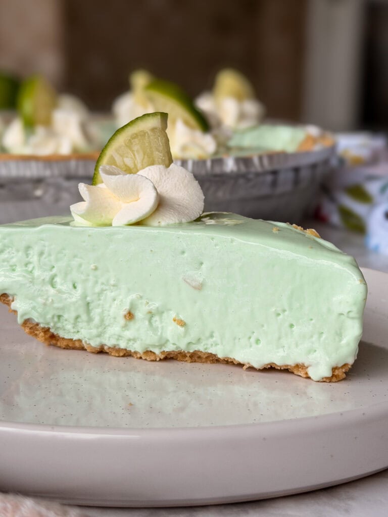 A slice of light green key lime pie with a graham cracker crust, topped with whipped cream and a lime wedge, sits on a white plate. The rest of the pie is visible in the background.