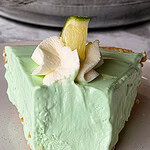 A slice of light green key lime pie with a crumbly graham cracker crust, topped with a swirl of whipped cream and a small wedge of lime, sits on a white plate.