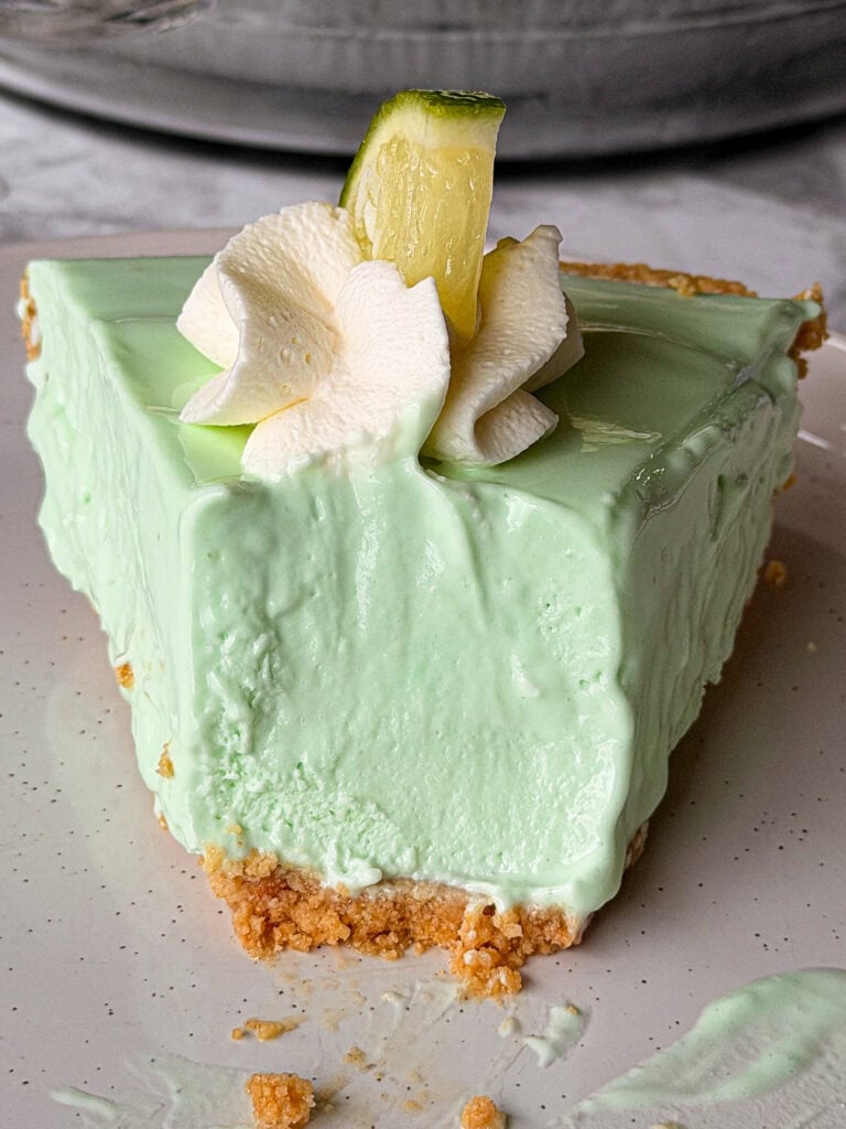 A slice of light green key lime pie with a crumbly graham cracker crust, topped with a swirl of whipped cream and a small wedge of lime, sits on a white plate.