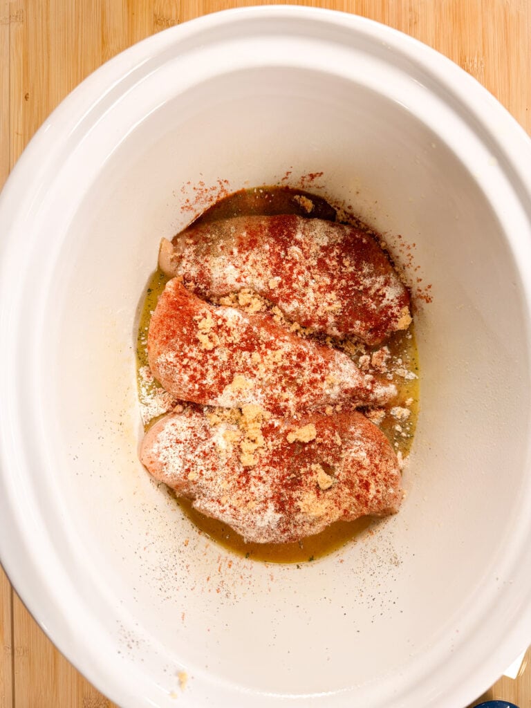 Three raw chicken breasts in a white slow cooker, topped with various seasonings including paprika, garlic powder, and grated parmesan, sitting in a layer of olive oil on a wooden surface.