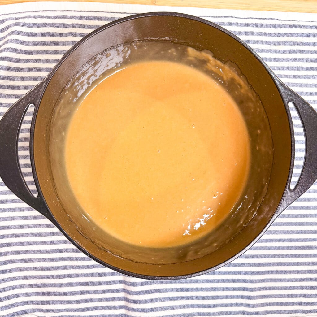 A cast iron pot filled with light brown roux sits on a white and gray striped cloth, viewed from above.