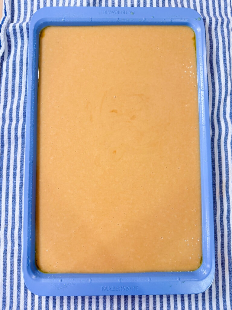 A blue rectangular baking pan filled with light tan cake batter sits on a blue-and-white striped cloth.