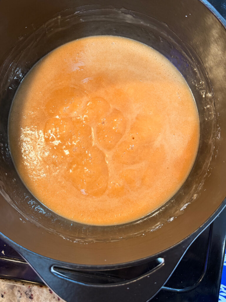 Orange, bubbling liquid cooking in a black pot on a stovetop, possibly a sauce or soup being prepared. The pot is partially visible, and the liquid appears to be in the process of simmering.