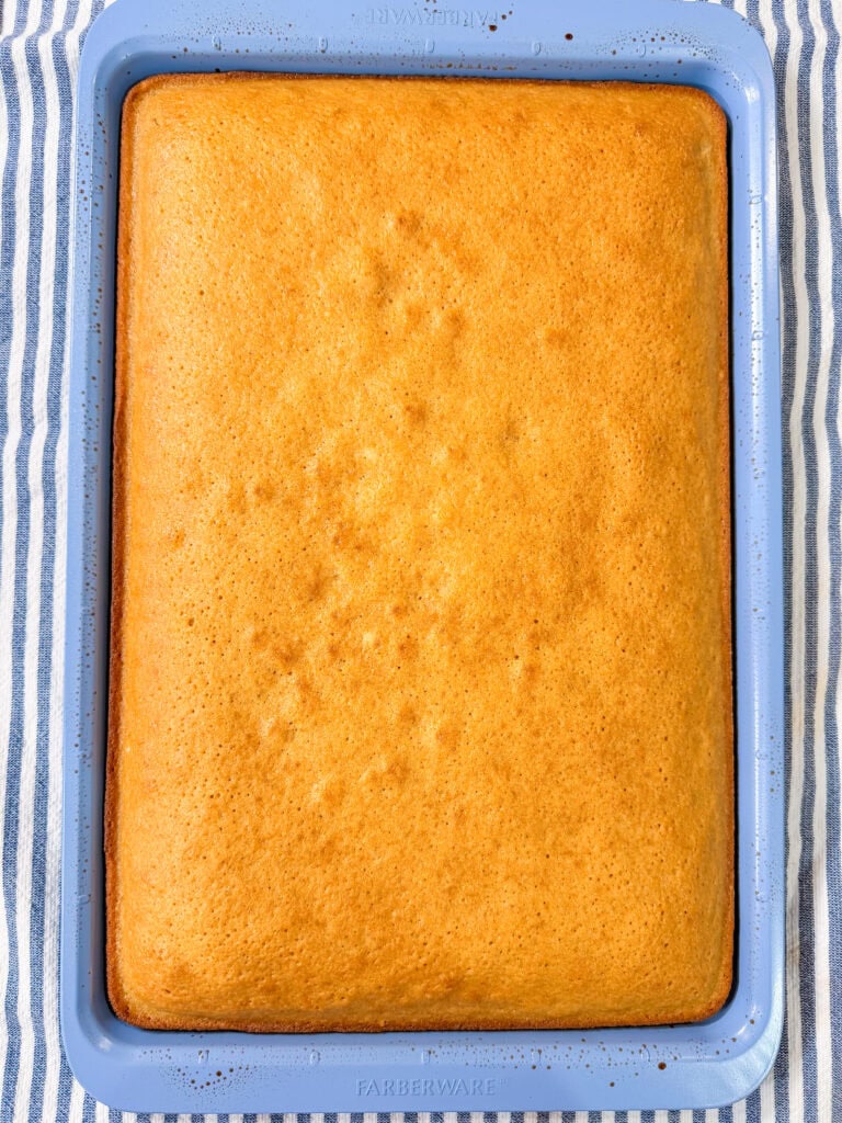 A golden-brown rectangular cake in a blue baking pan, resting on a blue and white striped cloth.