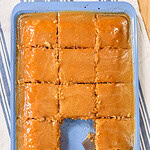 A rectangular cake in a blue baking pan, cut into 15 pieces with one piece missing. A spatula rests in the empty space, and caramel or butterscotch sauce covers the top and bottom.