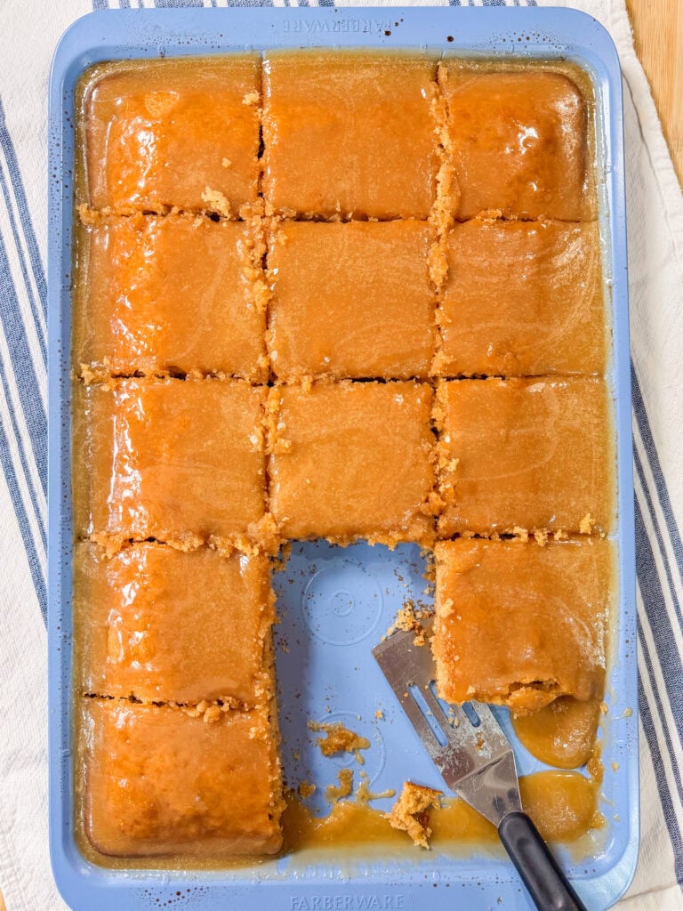 A rectangular cake in a blue baking pan, cut into 15 pieces with one piece missing. A spatula rests in the empty space, and caramel or butterscotch sauce covers the top and bottom.