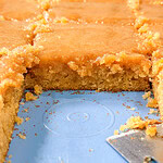A close-up of sliced, golden-brown cake with a glossy glaze on top. One piece is removed, revealing the soft, crumbly texture inside. A metal spatula rests on the blue tray. Crumbs are scattered around.