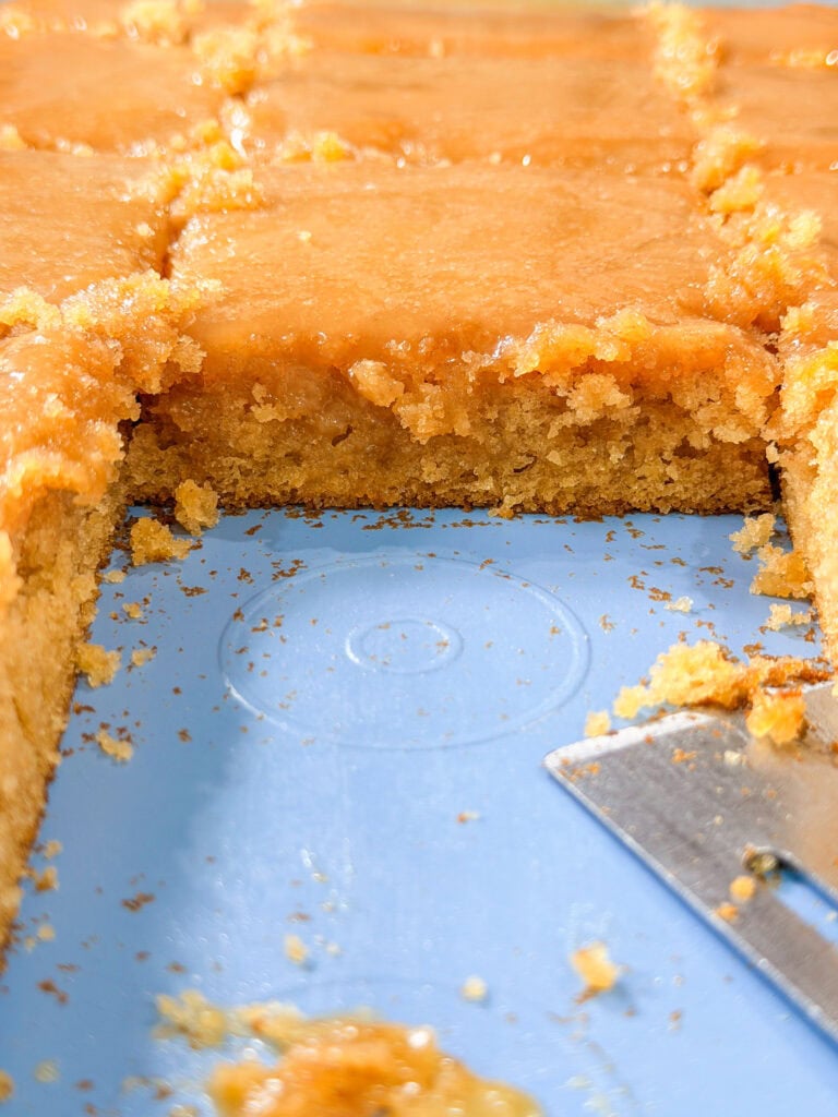 A close-up of sliced, golden-brown cake with a glossy glaze on top. One piece is removed, revealing the soft, crumbly texture inside. A metal spatula rests on the blue tray. Crumbs are scattered around.