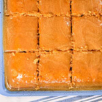 A tray of golden brown dessert cut into 12 square pieces, with a shiny, syrupy glaze on top. The tray rests on a white cloth with blue stripes.