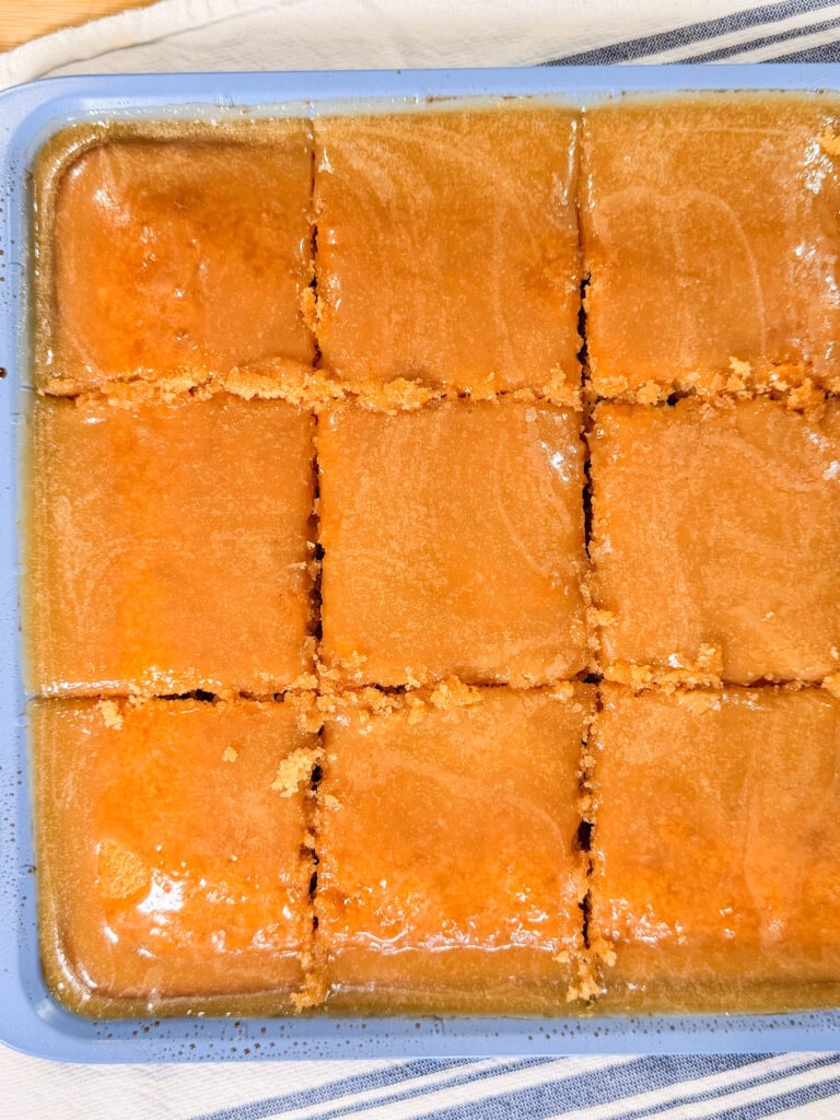 A tray of golden brown dessert cut into 12 square pieces, with a shiny, syrupy glaze on top. The tray rests on a white cloth with blue stripes.