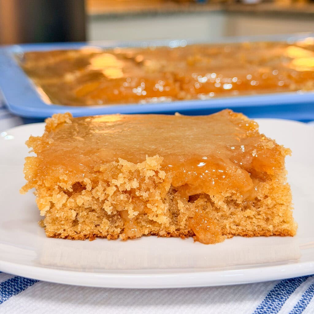 A close-up of a square piece of moist, golden brown cake with a gooey caramel-like topping on a white plate, with the rest of the cake in a baking pan in the background.