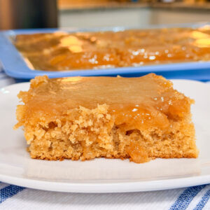 A close-up of a square piece of moist, golden brown cake with a gooey caramel-like topping on a white plate, with the rest of the cake in a baking pan in the background.