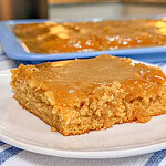 A close-up of a moist, golden-brown cake square with a caramel glaze on top sits on a white plate. In the background, more of the same cake is visible in a blue baking tray on a kitchen counter.
