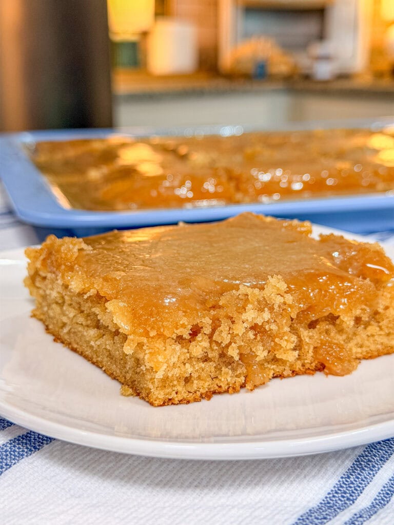 A close-up of a moist, golden-brown cake square with a caramel glaze on top sits on a white plate. In the background, more of the same cake is visible in a blue baking tray on a kitchen counter.