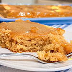 A close-up of a moist, golden brown cake with caramel glaze on a white plate and a fork, with a pan of the same cake in the background.