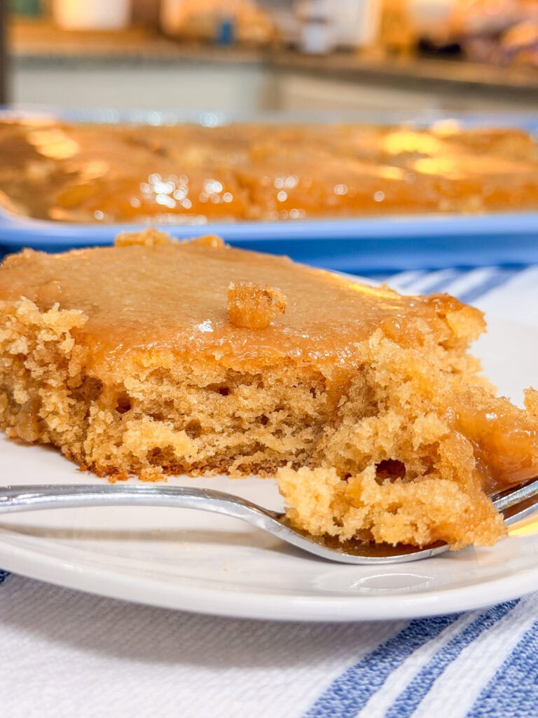 A close-up of a moist, golden brown cake with caramel glaze on a white plate and a fork, with a pan of the same cake in the background.
