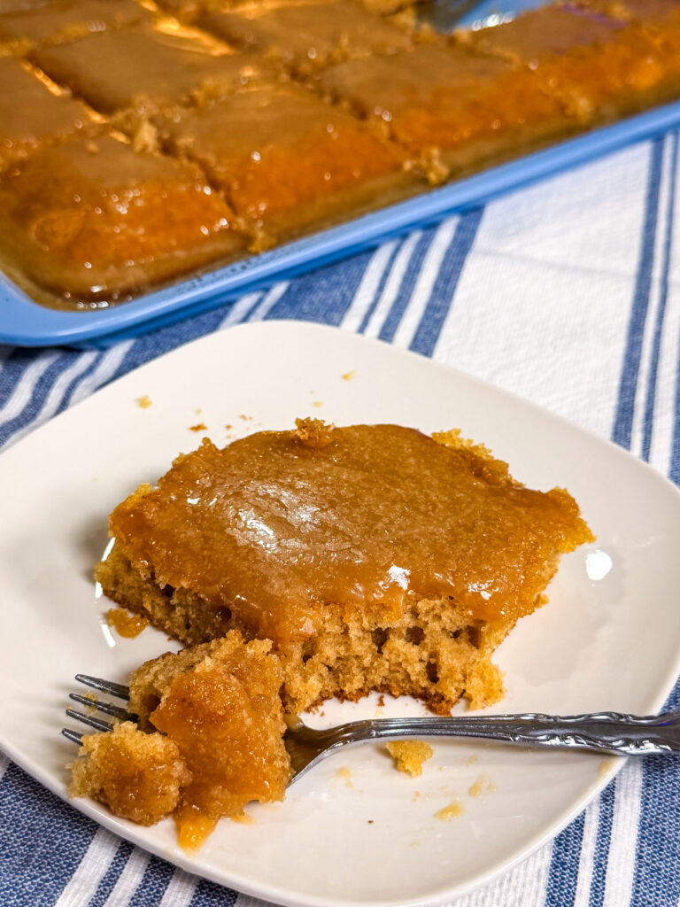 A slice of golden, moist cake on a white plate with a fork. The plate sits on a striped cloth, and a pan of more cake is visible in the background.