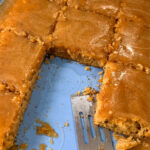 A blue baking pan with caramel-glazed sheet cake cut into squares. One square is missing, and a metal spatula rests in the empty space, with crumbs and caramel sauce left behind.