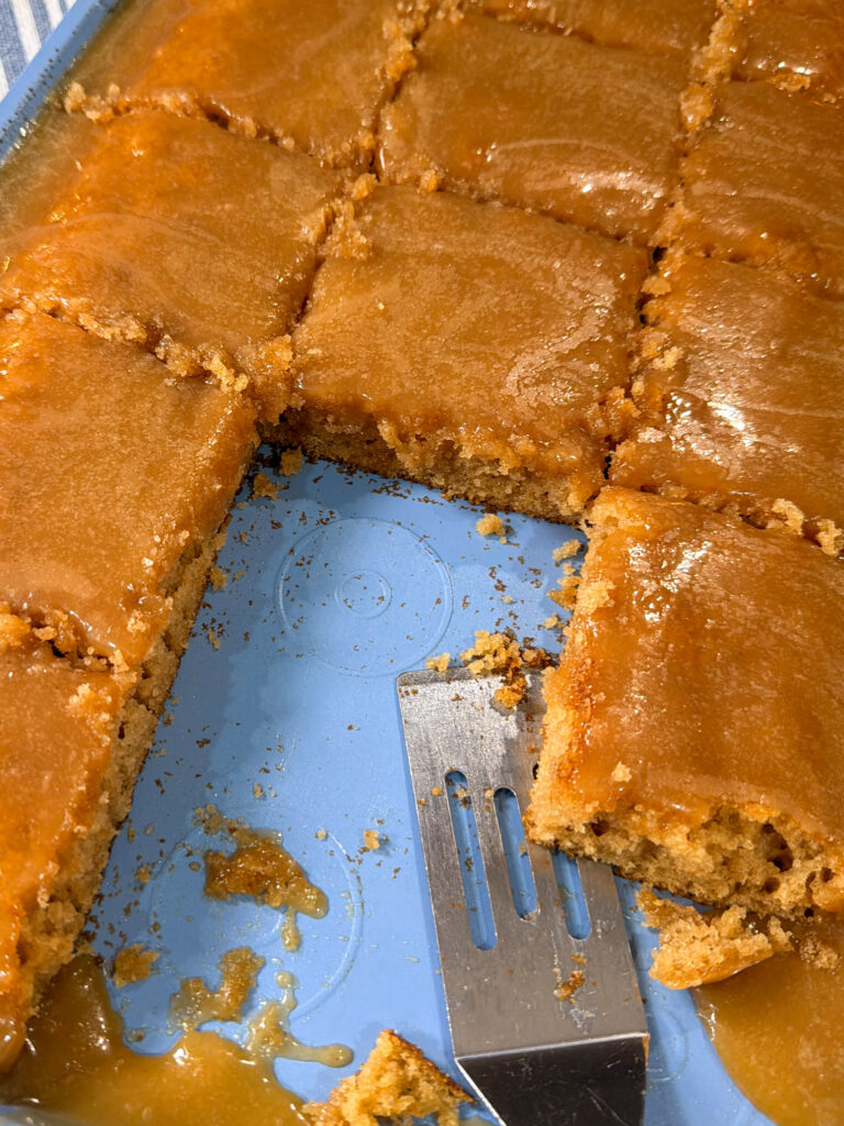 A blue baking pan with caramel-glazed sheet cake cut into squares. One square is missing, and a metal spatula rests in the empty space, with crumbs and caramel sauce left behind.