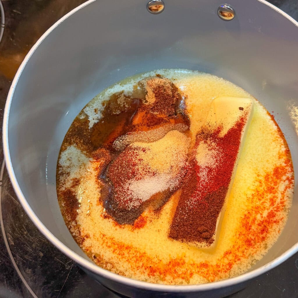 A saucepan with melted butter, a stick of unmelted butter, and various spices, including paprika and garlic powder, on a stovetop, beginning to blend together.