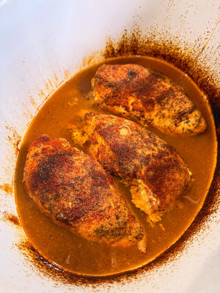 Three seasoned chicken breasts are cooking in a slow cooker, surrounded by a rich, orange-brown sauce with visible spices and juices. The sides of the slow cooker show some sauce splatter.