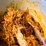 Tongs holding up a portion of shredded, seasoned meat over a bowl filled with more of the same meat, likely freshly cooked and juicy with visible spices.