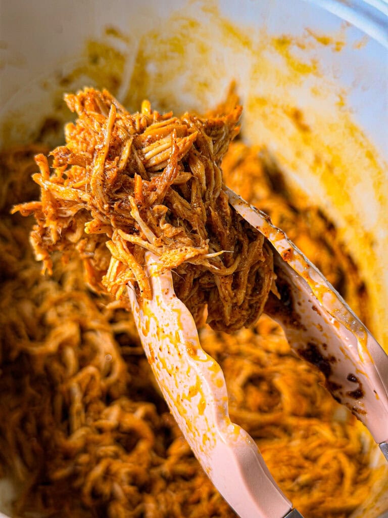 Tongs holding up a portion of shredded, seasoned meat over a bowl filled with more of the same meat, likely freshly cooked and juicy with visible spices.