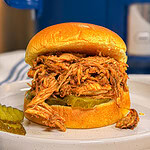 A shredded barbecue chicken sandwich on a toasted bun with pickle slices, served on a white plate. A few pickle slices and a small piece of chicken are beside the sandwich. A blue kitchen appliance is in the background.