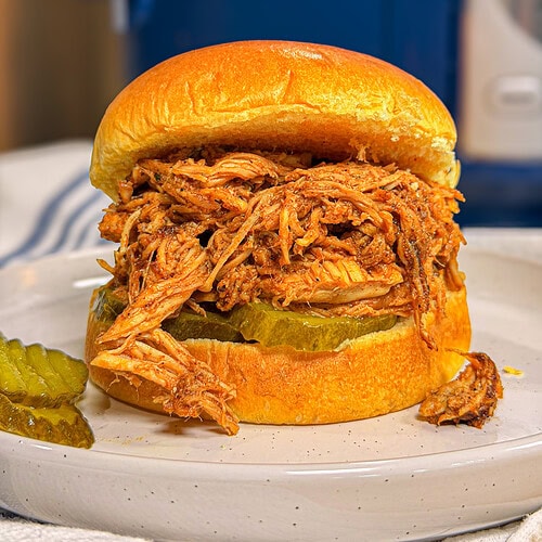 A shredded barbecue chicken sandwich on a toasted bun with pickle slices, served on a white plate. A few pickle slices and a small piece of chicken are beside the sandwich. A blue kitchen appliance is in the background.