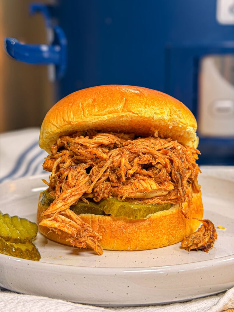 A shredded barbecue chicken sandwich on a toasted bun with pickle slices, served on a white plate. A few pickle slices and a small piece of chicken are beside the sandwich. A blue kitchen appliance is in the background.