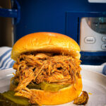 A pulled pork sandwich with pickles on a white plate, in front of a blue Crock-Pot slow cooker. The sandwich is on a soft bun, and extra pickle slices are on the plate.