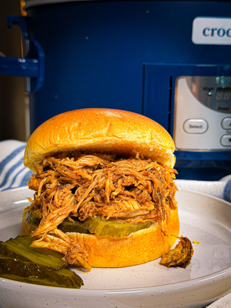 A pulled pork sandwich with pickles on a white plate, in front of a blue Crock-Pot slow cooker. The sandwich is on a soft bun, and extra pickle slices are on the plate.