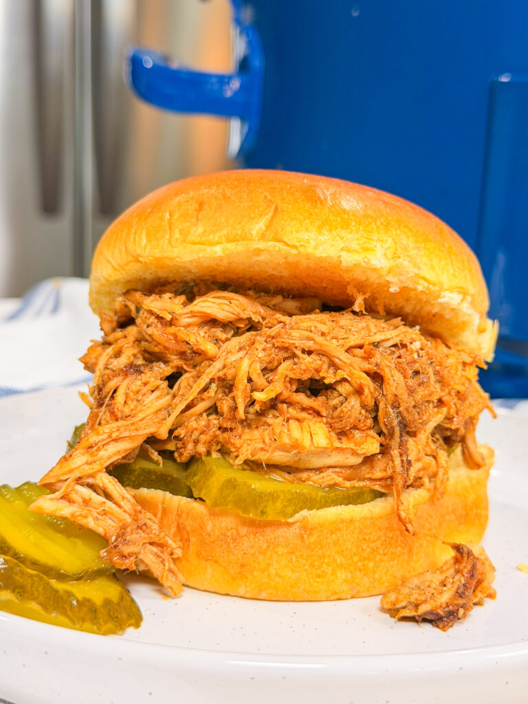 A close-up of a pulled barbecue chicken sandwich on a brioche bun with pickle slices, served on a white plate. A few extra pickles and a blue pot are visible in the background.