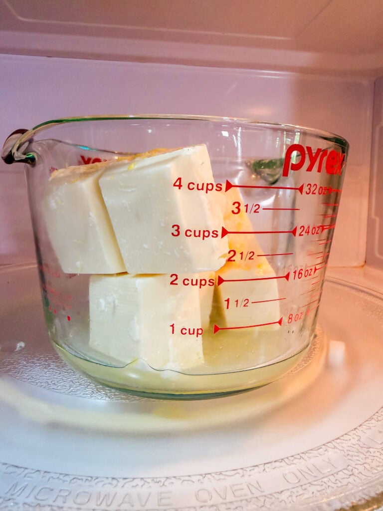 A glass measuring cup with cubes of butter inside sits on a microwave turntable. The cup is marked with red measurement lines up to 4 cups.