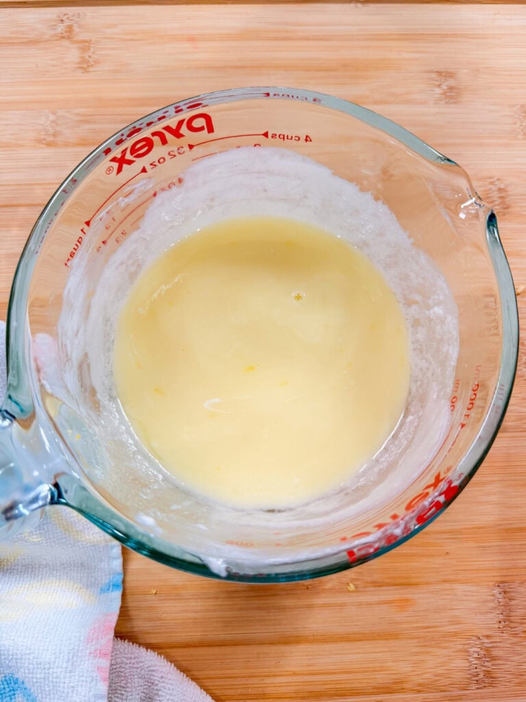 A glass measuring cup with a light yellow batter or mixture inside, placed on a wooden surface. A towel is partially visible on the left side of the image.