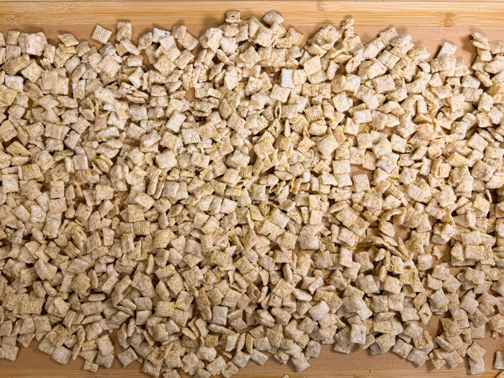 A large pile of square, lightly textured breakfast cereal pieces spread out evenly on a wooden surface.