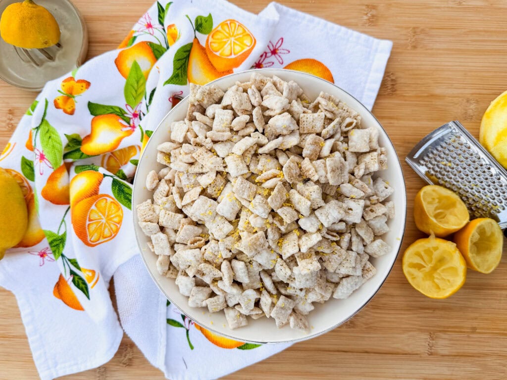 A bowl of powdered cereal snack sits on a wooden surface, surrounded by lemon halves, a grater, a citrus-themed towel, and a citrus juicer.