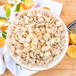 A bowl of powdered sugar-coated cereal squares sits on a wooden surface beside a citrus-themed towel, a grater, and sliced lemons.