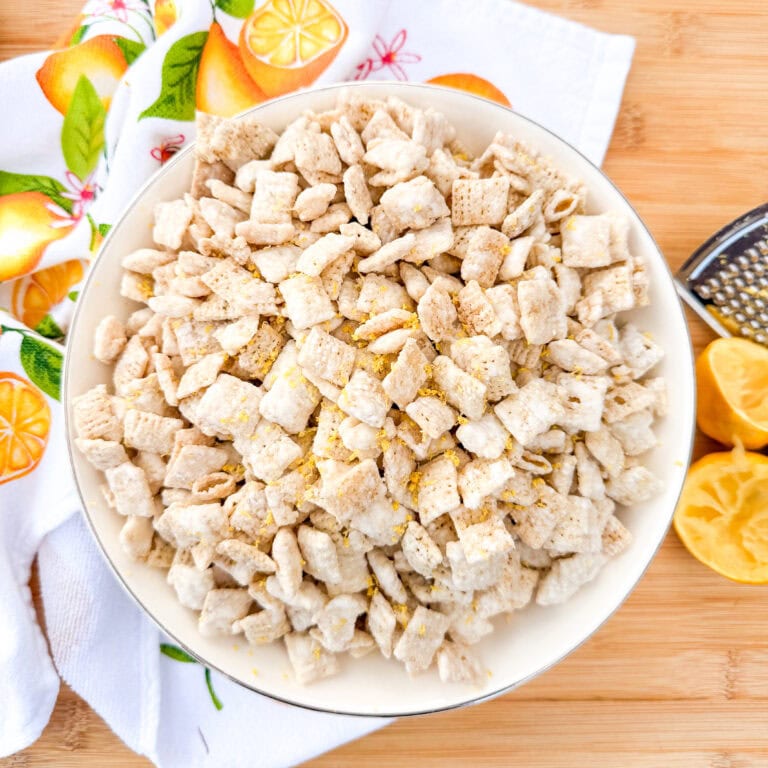 A bowl of powdered sugar-coated cereal squares sits on a wooden surface beside a citrus-themed towel, a grater, and sliced lemons.