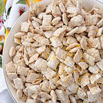 A bowl filled with powdered sugar-coated cereal squares, possibly puppy chow or muddy buddies, sprinkled with yellow zest, placed on a white towel with a colorful fruit pattern.