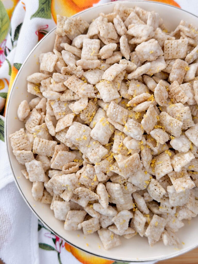 A bowl filled with powdered sugar-coated cereal squares, possibly puppy chow or muddy buddies, sprinkled with yellow zest, placed on a white towel with a colorful fruit pattern.