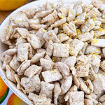 A bowl of lemon-flavored snack mix coated in powdered sugar sits on a table with whole and halved lemons, a grater, and a citrus zester in the background.