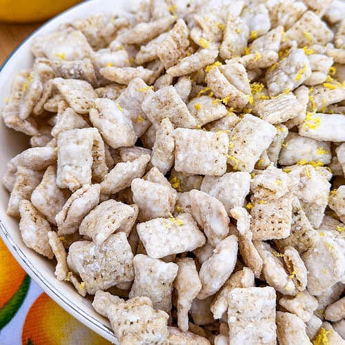 A close-up of a bowl filled with powdered sugar-coated cereal squares, sprinkled with lemon zest. Whole and sliced lemons and a grater are visible in the blurred background.