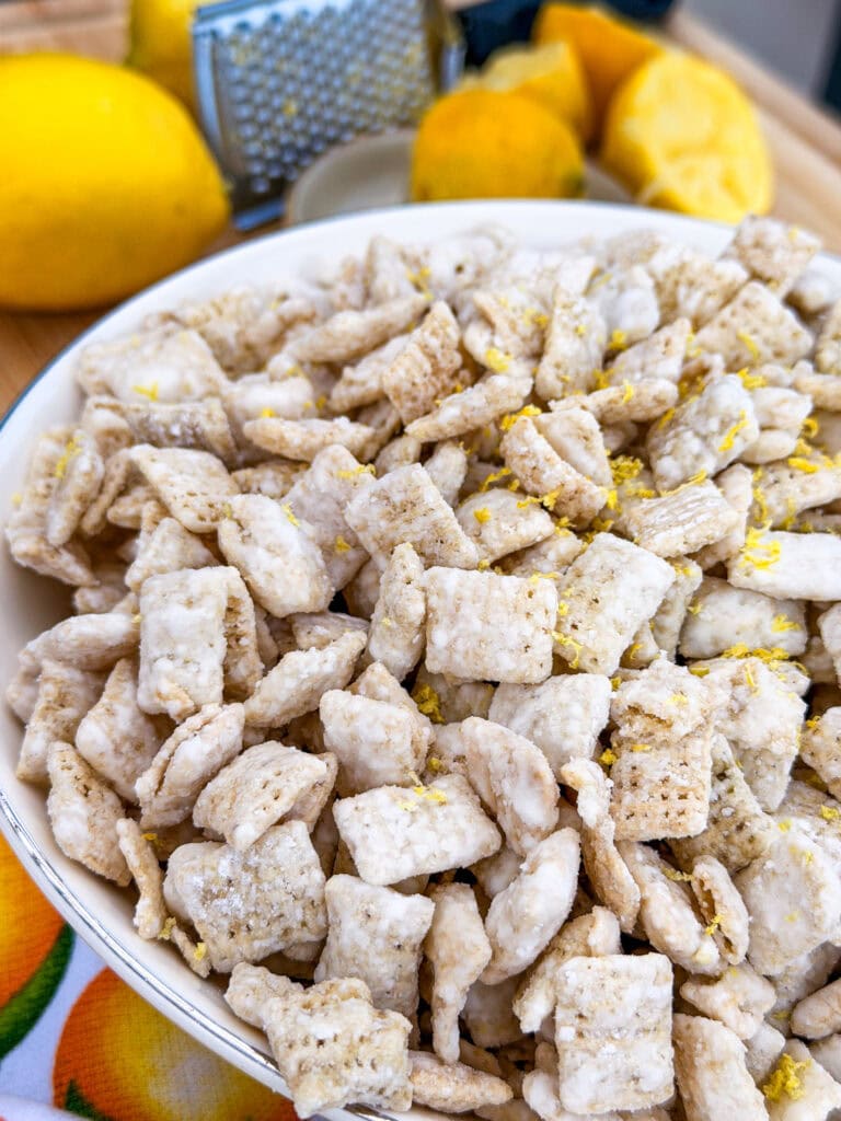 A close-up of a bowl filled with powdered sugar-coated cereal squares, sprinkled with lemon zest. Whole and sliced lemons and a grater are visible in the blurred background.