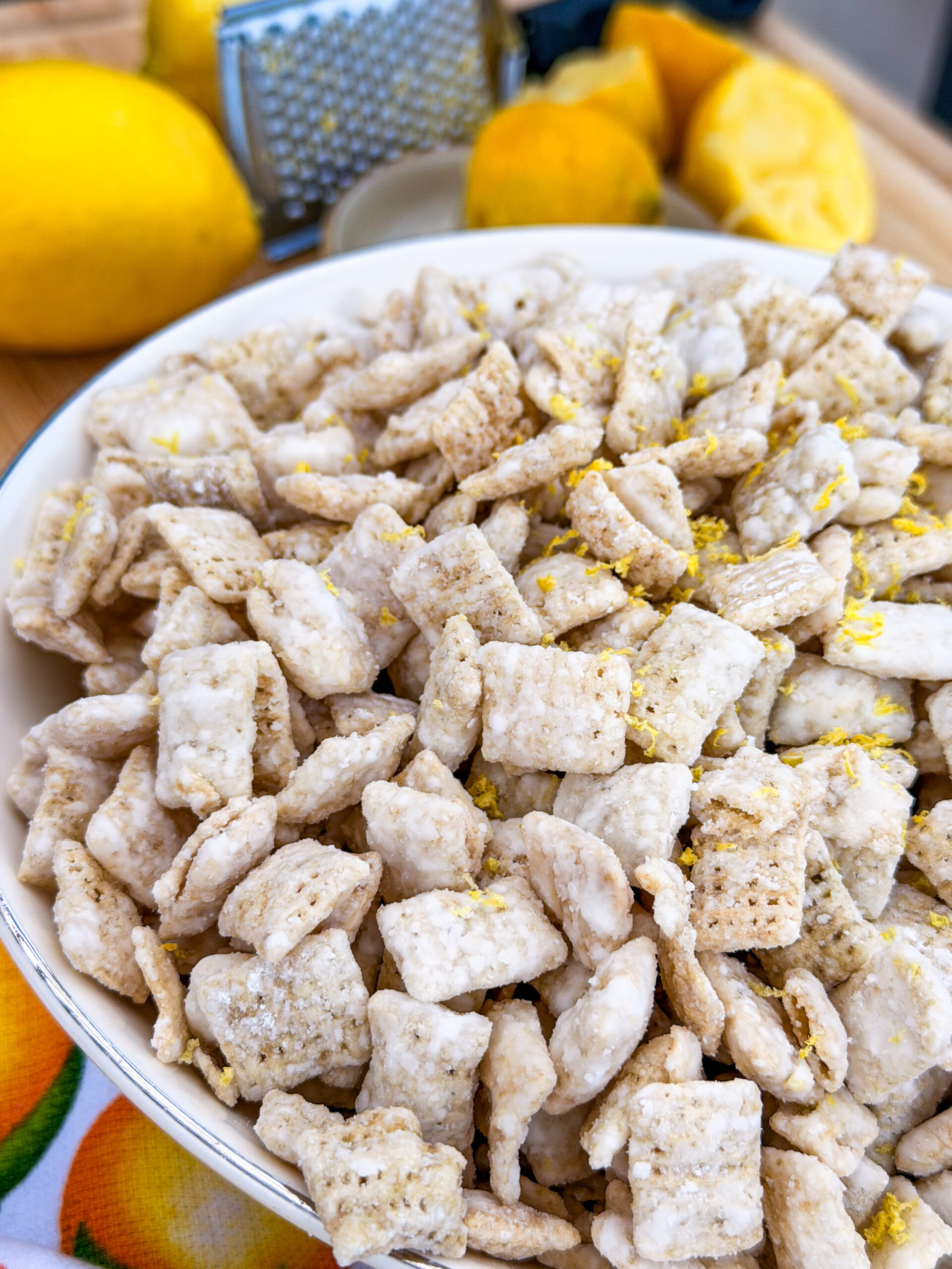 A close-up of a bowl filled with powdered sugar-coated cereal squares, sprinkled with lemon zest. Whole and sliced lemons and a grater are visible in the blurred background.